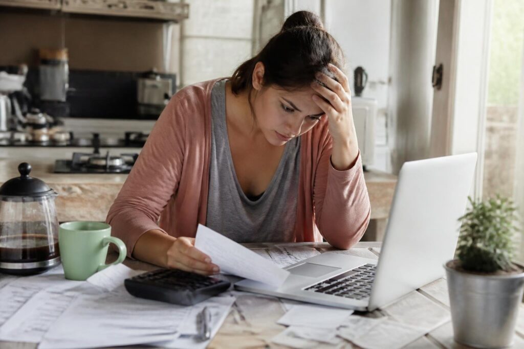 A young adult sitting at a kitchen table surrounded by bills and a calculator, looking thoughtful and concerned.
