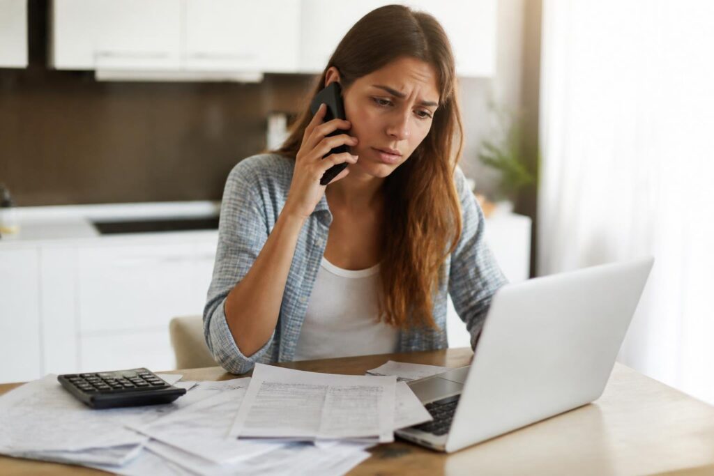 A worried young adult sitting at a kitchen table, holding a smartphone to their ear with bills and a laptop on the table.